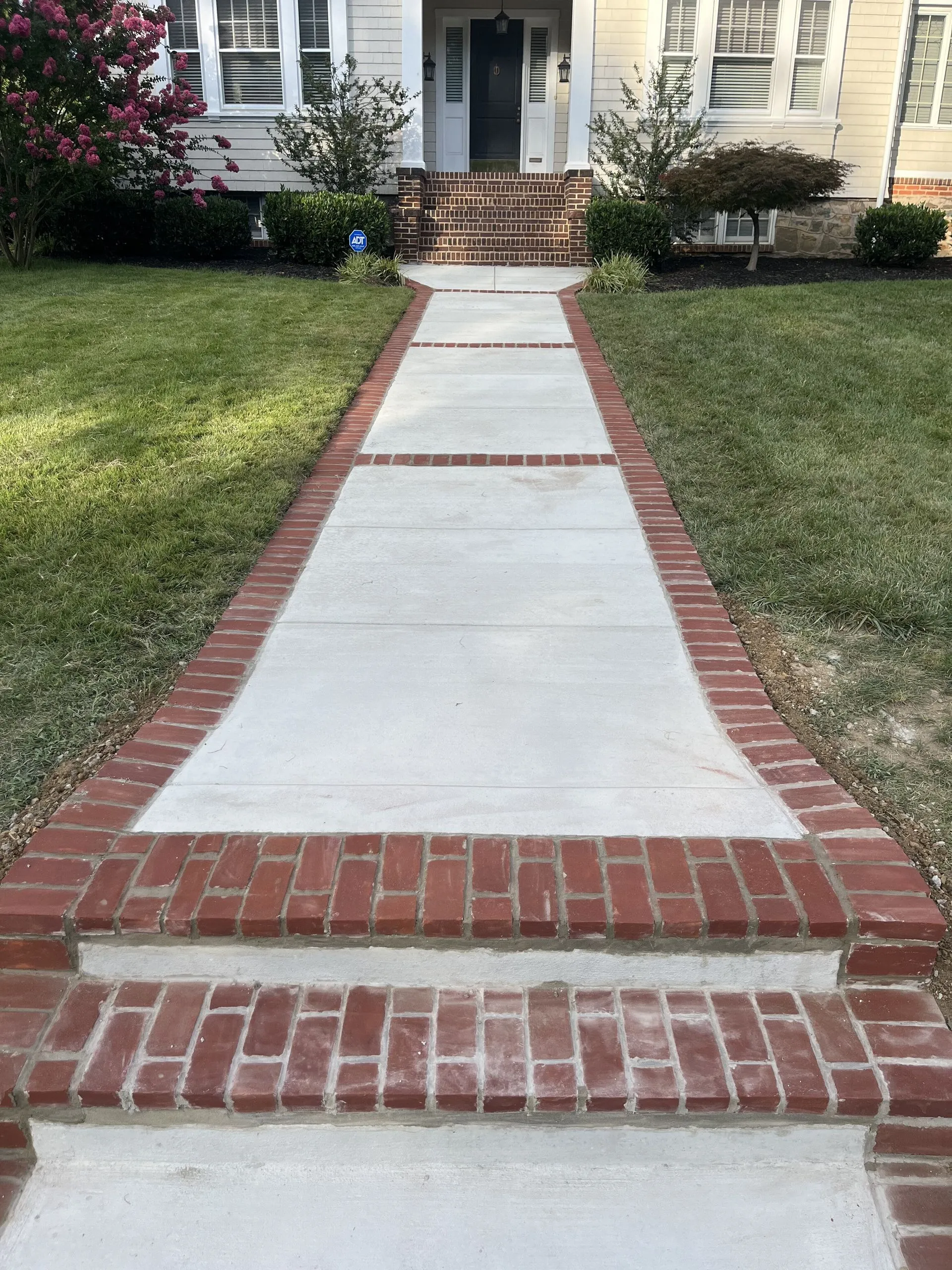 Concrete walkway with red brick borders and steps leading up to a light-colored house with a dark front door