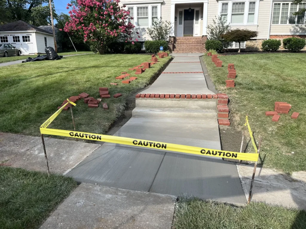 A freshly poured concrete walkway leading to a house entrance, cordoned off with yellow caution tape, with red bricks staged along both sides for a decorative border.