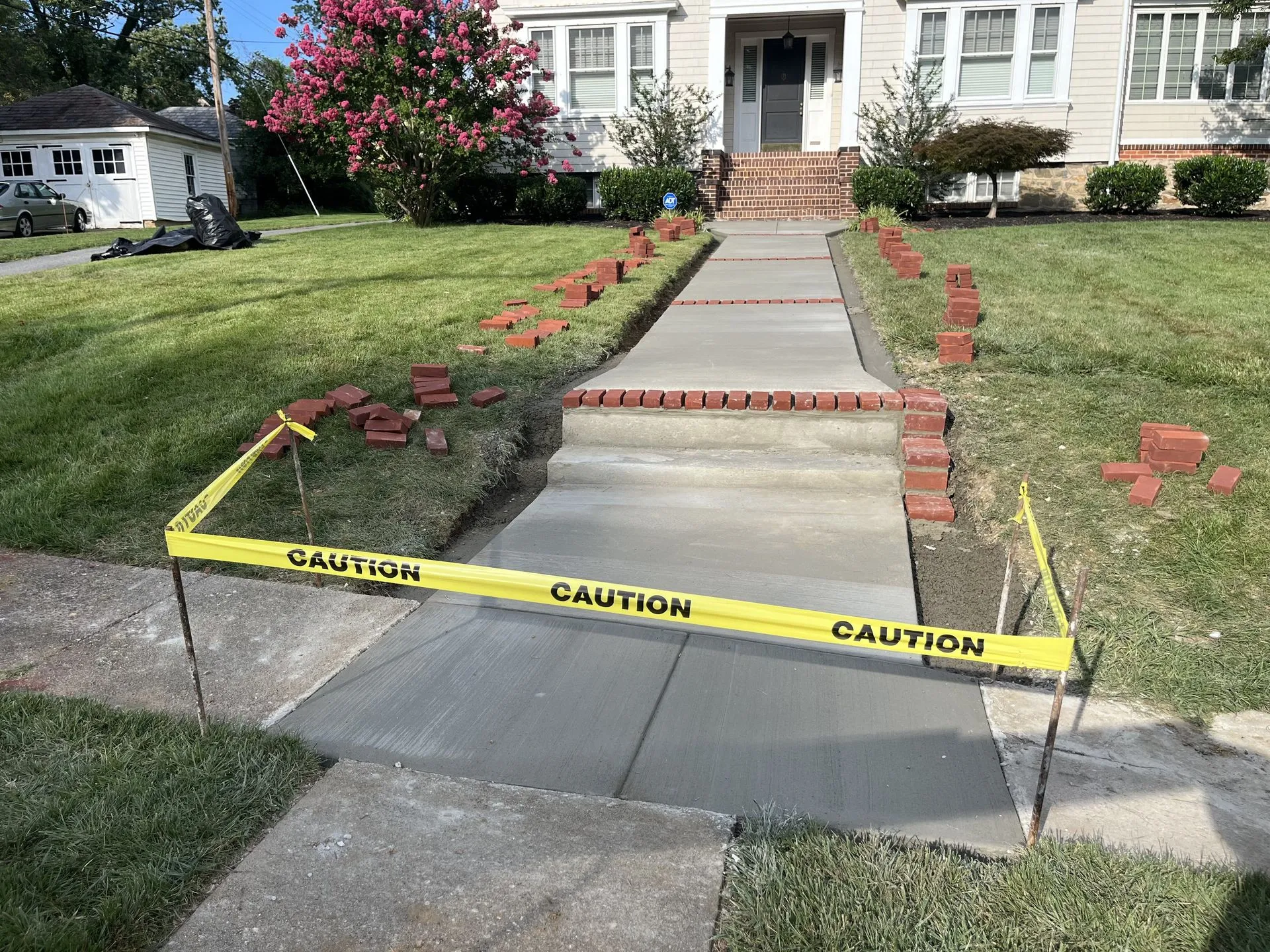 Freshly poured concrete walkway leading to a house entrance, cordoned off with caution tape while curing, with brick staged for a decorative border