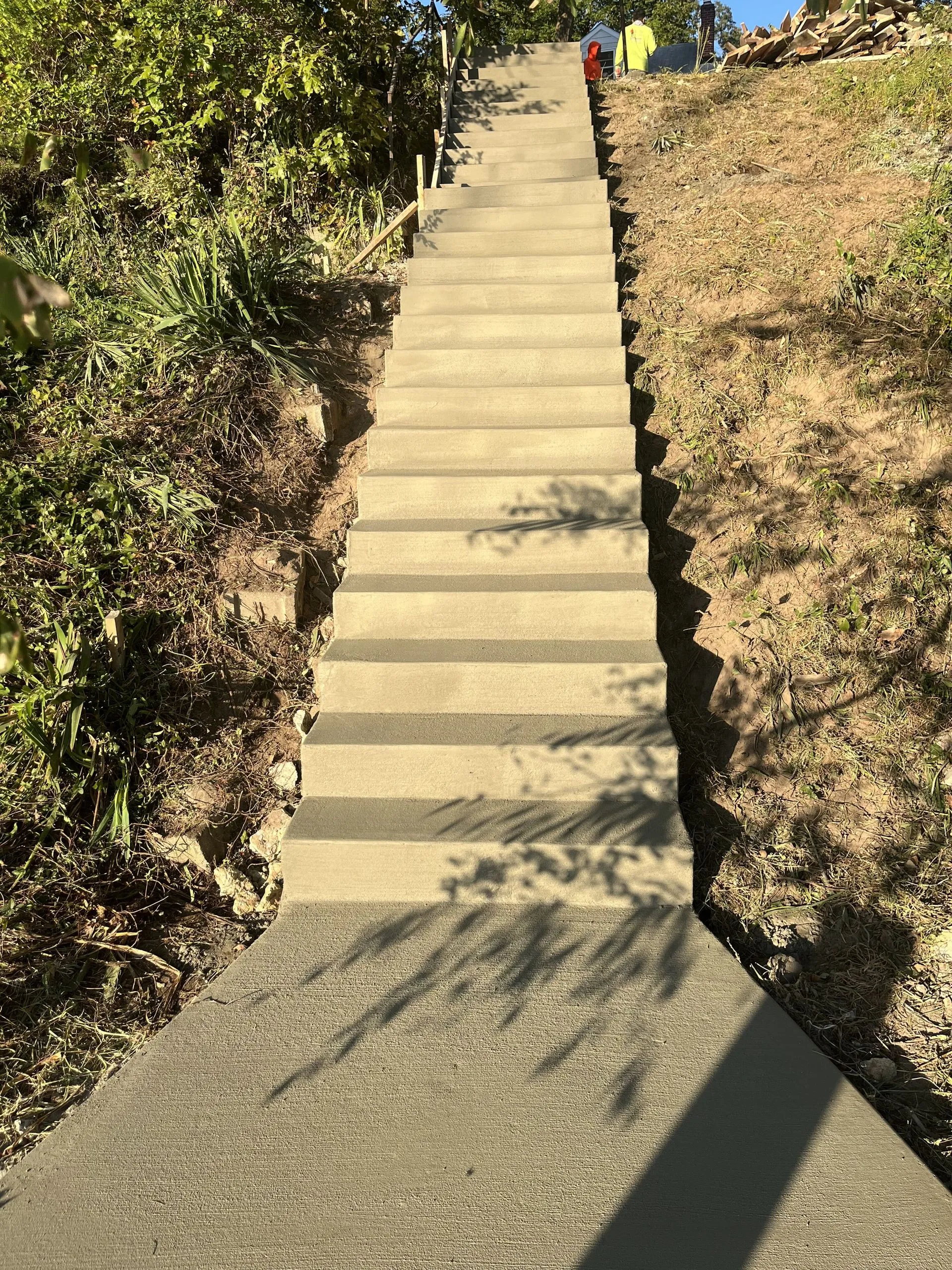 Long concrete staircase ascending a hillside between overgrown vegetation, with the crew working at the top