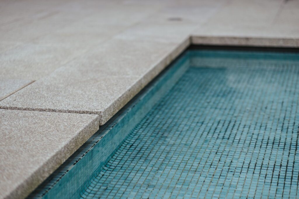 A close-up of a swimming pool corner with light stone paving edging and turquoise mosaic tile visible beneath the clear water.